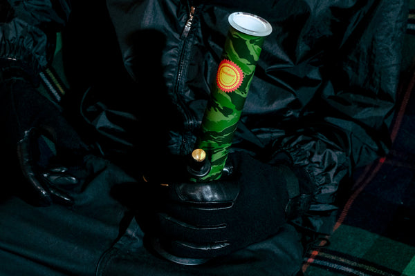 A seated man holding the Bong Warehouse Large Tube 33 cm Glass Bong in Camo Sunbaked pattern