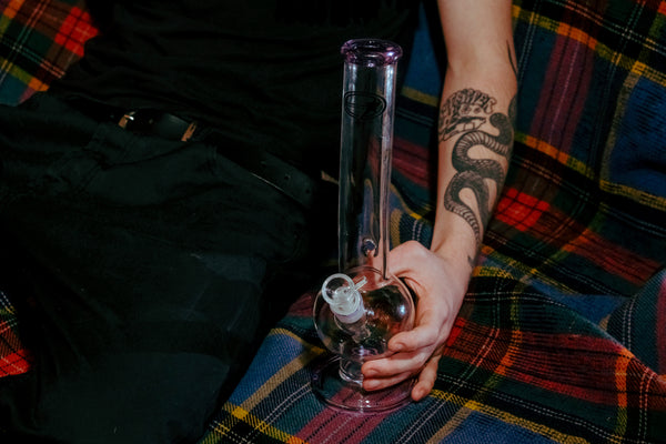 A stoner sitting while holding the Shotties™ 33 cm Glass Bent Bubble Bong in clear and purple glass from Bong Warehouse beside him