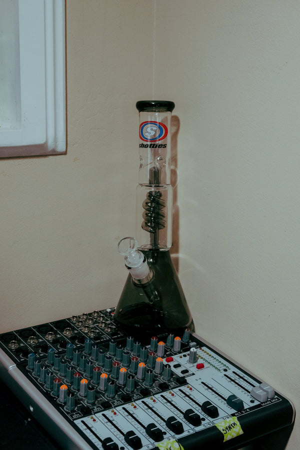 Shotties™ 30 cm Spiral Perk Glass Beaker Bong in clear and smokey glass from Bong Warehouse, displayed on top of a sound mixing board