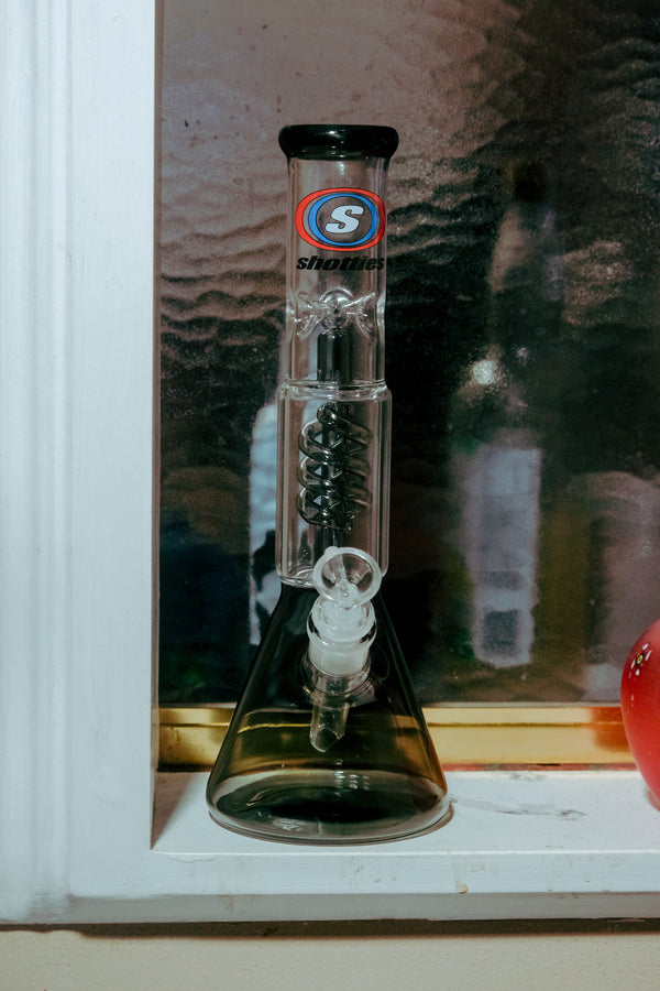 Shotties™ 30 cm Spiral Perk Glass Beaker Bong in clear smokey glass from Bong Warehouse, displayed on a windowsill
