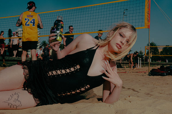 Woman posing seductively holding Bong Warehouse Black High Tiger 17.5cm Bottle Bong, with volleyball game in the background