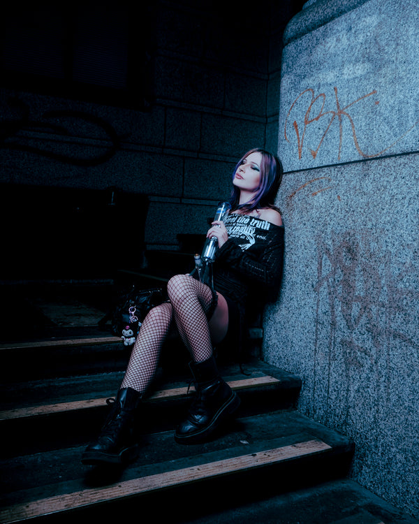 Seated on the stairs, holding a Shotties™ 33 cm Glass Perc Pillar Bong in clear and blue glass from Bong Warehouse