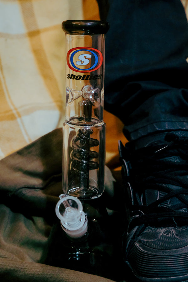 Shotties™ 30 cm Spiral Perk Glass Beaker Bong in clear and smokey glass from Bong Warehouse, placed beside a man's shoes
